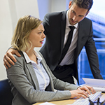 Workplace Harassment_Squareimage A female professional working on the computer reacts to workplace harassment by her colleague who places his hand over her shoulders.